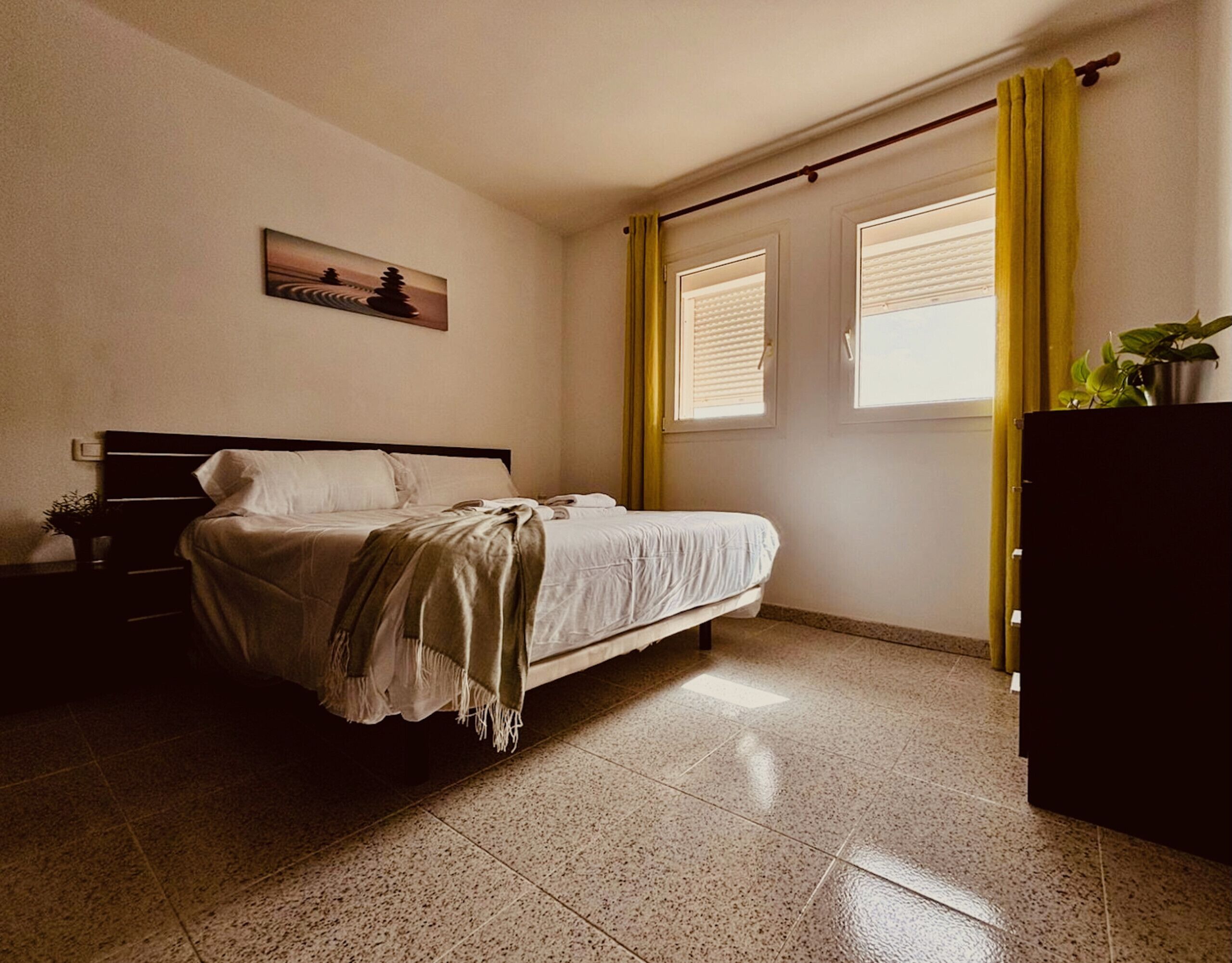 Une chambre à coucher avec un lit, une table de chevet, une fenêtre avec des stores, un rideau et une plante posée sur une commode.