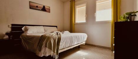 Une chambre à coucher avec un lit, une table de chevet, une fenêtre avec des stores, un rideau et une plante posée sur une commode.