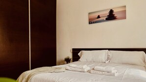 Una cama bien tendida con sábanas blancas y almohada, una pequeña planta en maceta en la mesita de noche, y un cuadro de un paisaje montañoso en la pared.