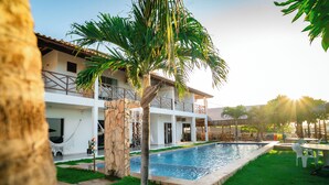 A pool area with a palm tree, a stone wall, and a white building with balconies. - Milano Hotel Pousada Canoa Quebrada (Aracati)