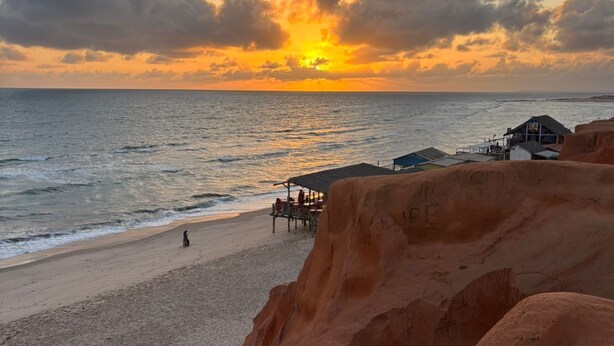 A beach with a sunset, rocky cliffs, and a small building with a roof. - Milano Hotel Pousada Canoa Quebrada (Aracati)