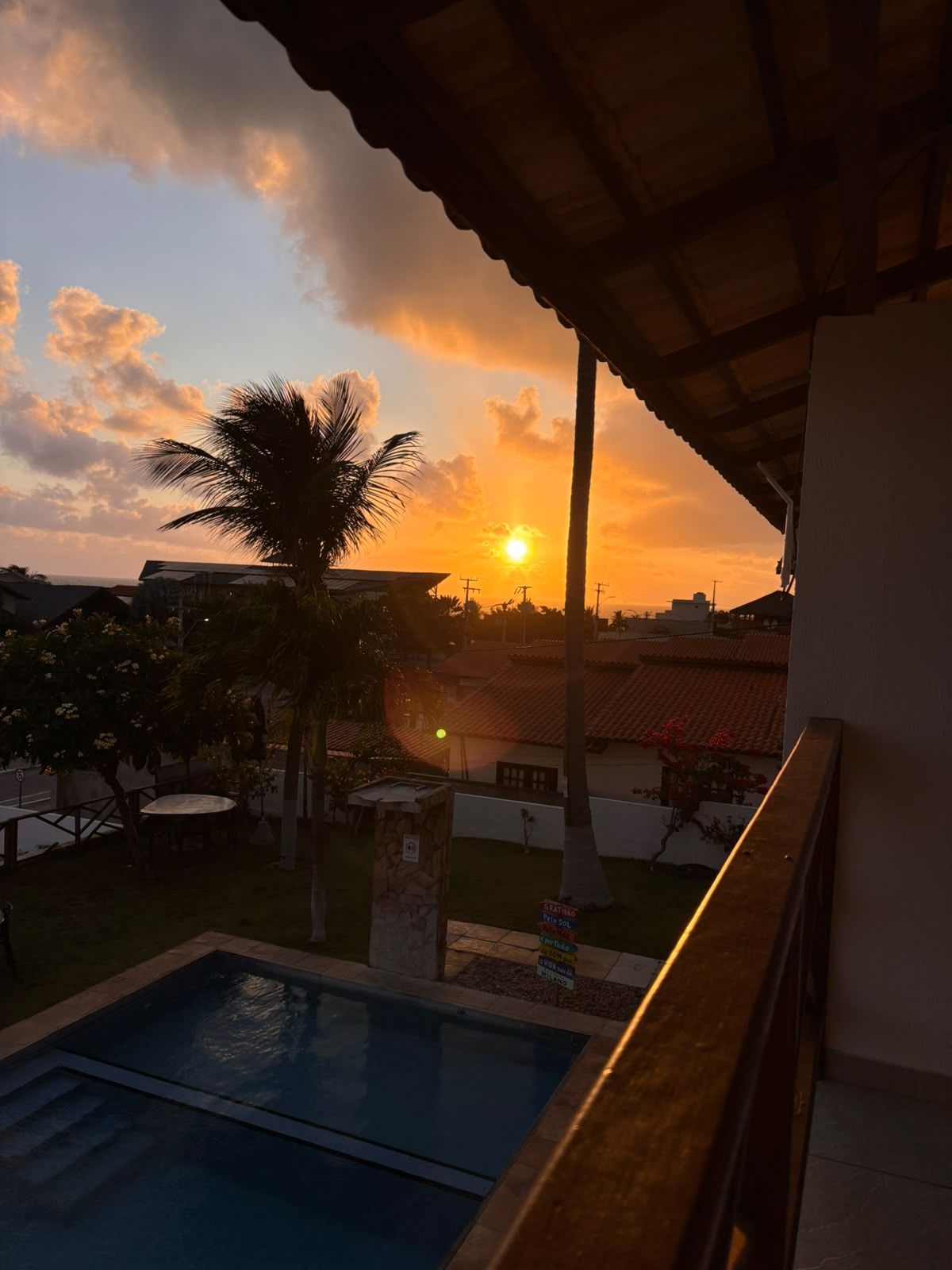 A sunset view from a covered balcony overlooking a swimming pool and a residential area.