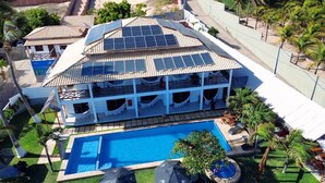 A large building with a swimming pool, solar panels on the roof, and a fenced yard with palm trees. - Milano Hotel Pousada Canoa Quebrada (Aracati)
