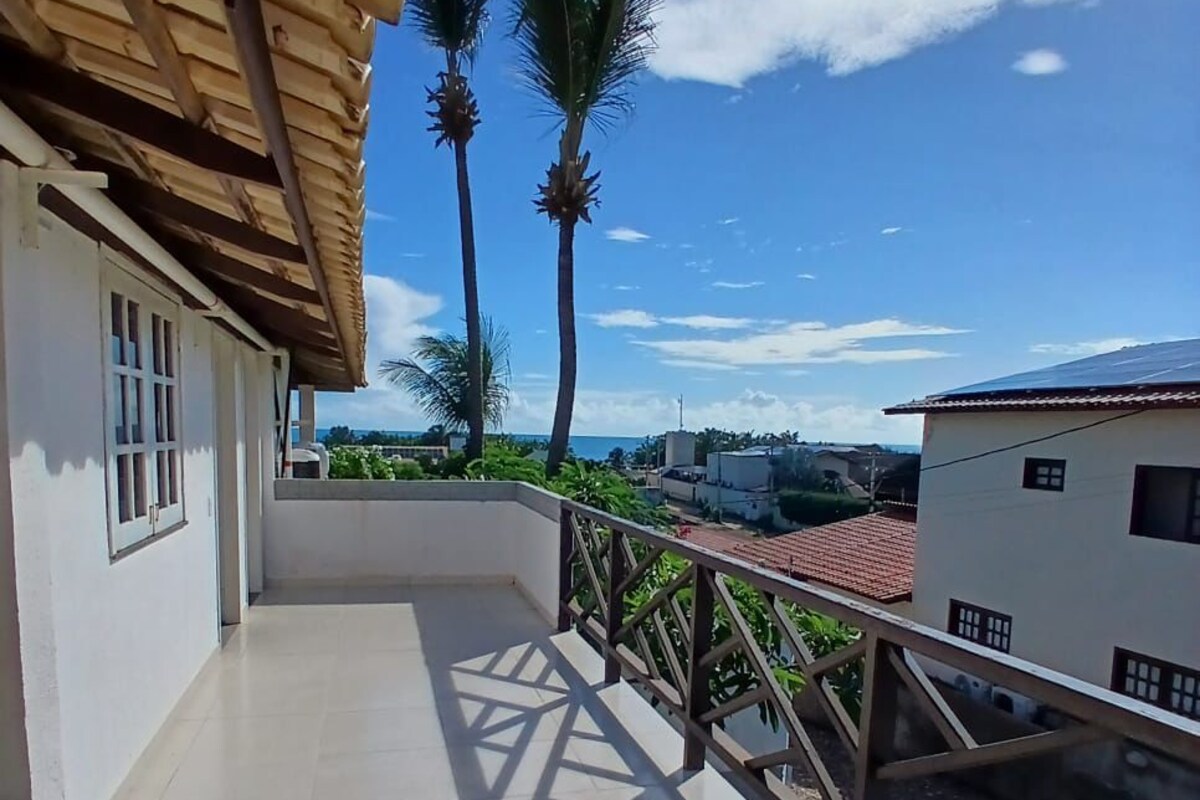 A balcony with a tiled floor, a wooden railing, and a view of palm trees and buildings in the distance.