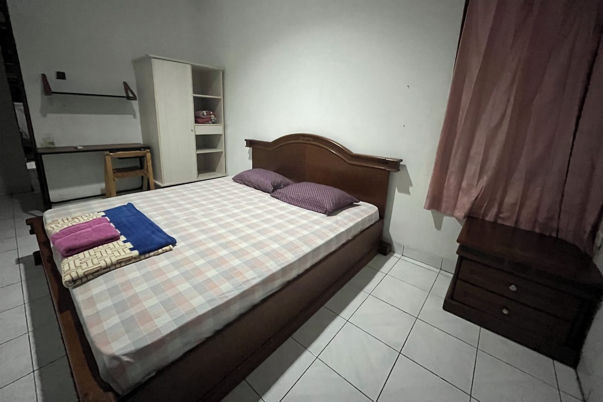 Un lit simple avec une couvre-lit à carreaux, une tête de lit en bois et deux oreillers. Il y a une armoire avec des étagères ouvertes sur la gauche et une petite table avec une chaise. La pièce est dotée d'un sol carrelé blanc et d'un rideau rose sur la droite.