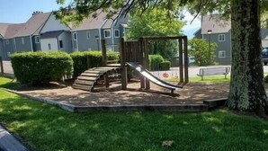 Ein Spielplatz mit Rutsche und Kletterstufen, umgeben von Gras und Bäumen, mit Häusern im Hintergrund.