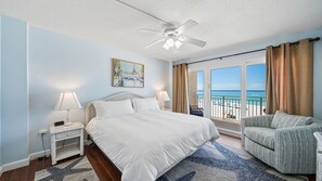 A hotel room with a bed, two bedside tables, a chair, a ceiling fan, a painting, and a view of the beach.