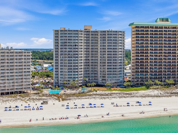 A beachfront hotel with multiple high-rise buildings, a sandy beach with blue umbrellas, and a clear view of the ocean.