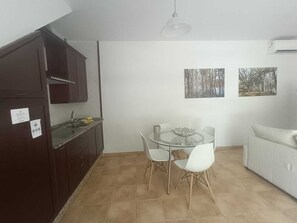 Cocina compacta con alacenas oscuras, un lavamanos y un comedor con mesa de vidrio y sillas blancas. Se observan dos cuadros colgados en la pared y un sofá blanco.