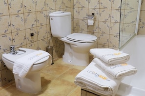 A bathroom with a toilet, bidet, and sink, all with white fixtures. There are towels with 'CostaSur' branding on the sink and stacked on the floor. - Hacienda Laguna Seca - Apart. Jazmin (Cadiz)