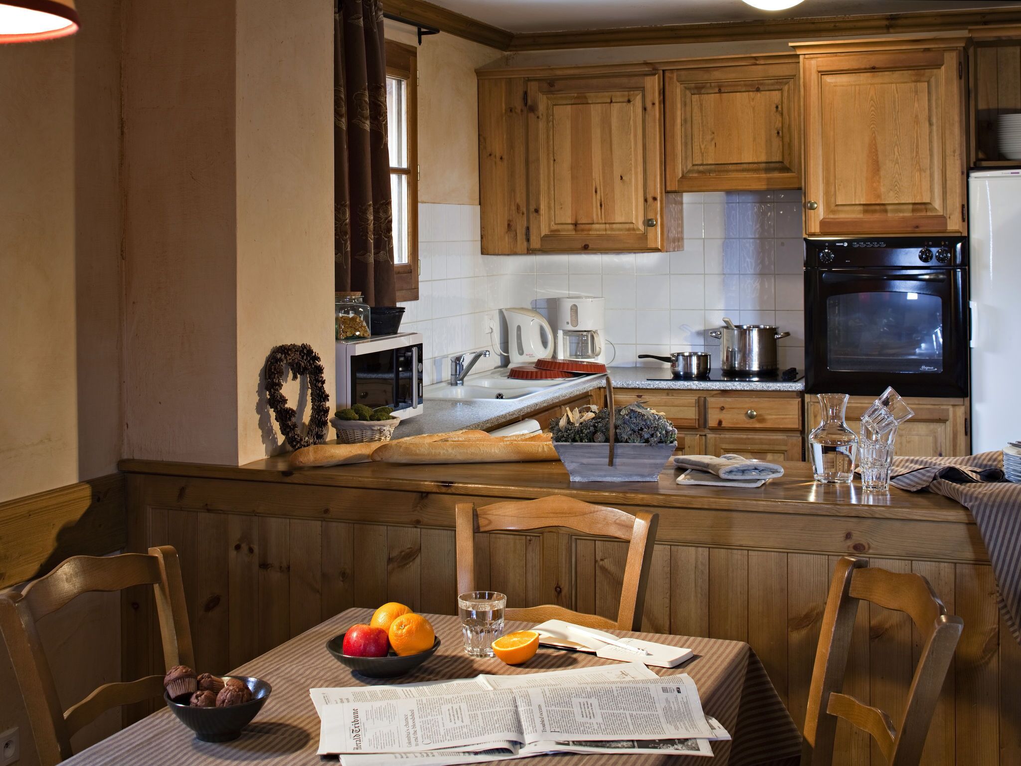 A kitchen with wooden cabinets, a stove, a coffee maker, and a dining table set with a bowl of fruit and a newspaper.