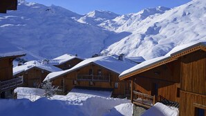 Uma fileira de cabanas de madeira com telhados cobertos de neve, ao fundo montanhas também cobertas de neve.
