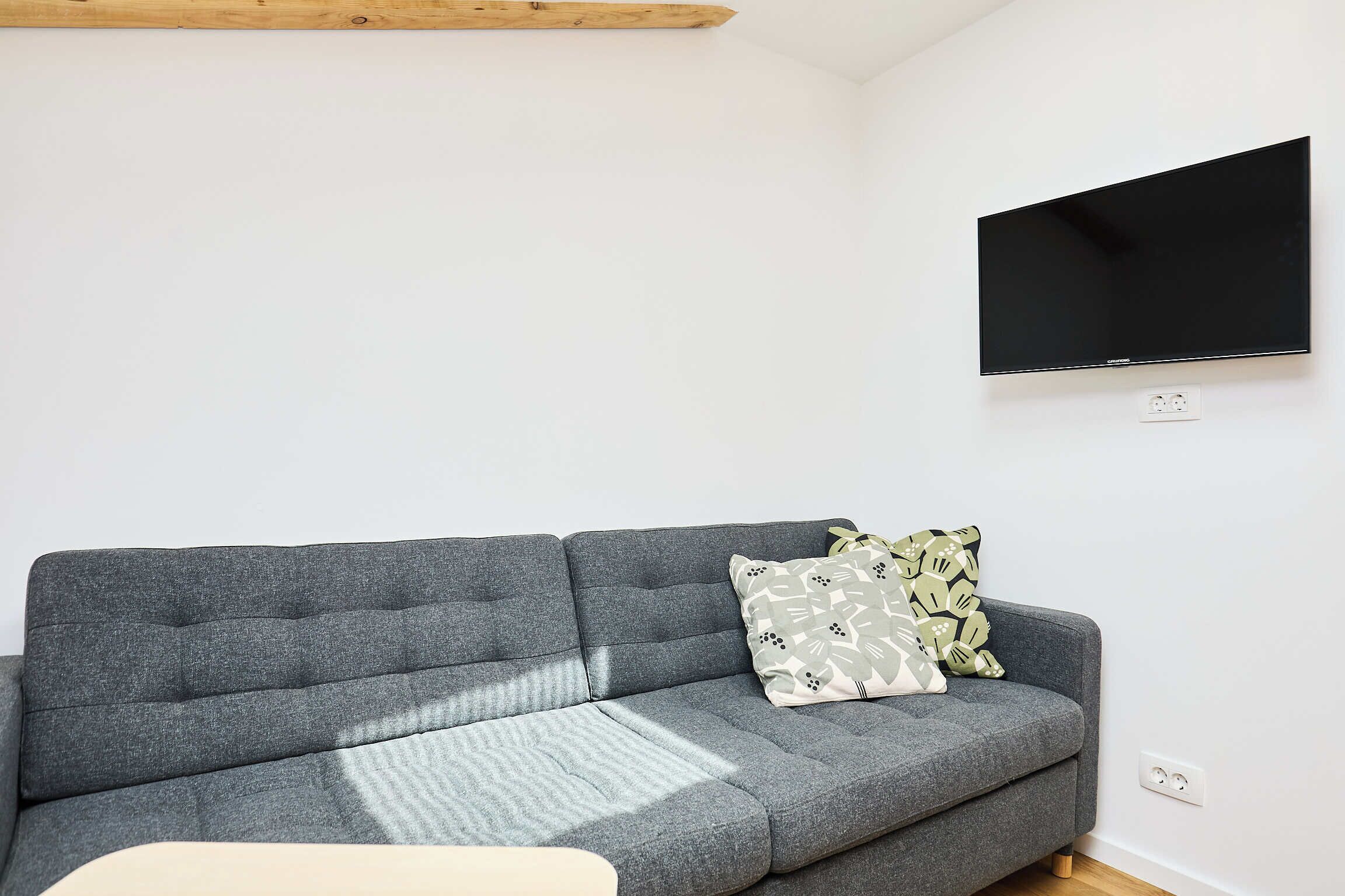 A modern living room with a grey sofa, patterned cushions, a mounted flat-screen TV, and a wooden beam ceiling.