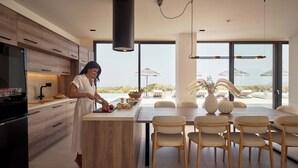 A modern kitchen with wooden cabinets and a large island. There is a dining area with chairs and a table, and a view of the outdoors through large windows. - Gavras Exclusive Villa II, Pool & Heated Whirlpool, SeaViews (9HFX+3FH Arkadi, Greece)