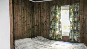 Una habitación con paredes de madera, una cama con colcha blanca y una ventana con cortinas coloridas.