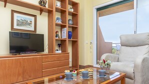A living room with a wooden TV stand, a glass coffee table, a beige armchair, and a bookshelf with various decorative items. - Port Pals Sea View by Interhome (Pals)