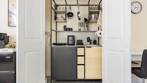 Cocina compacta con refrigerador, estufa y pileta. Hay una pequeña mesada con cafetera y tetera. La cocina tiene alacenas de madera y un colgador para ollas.