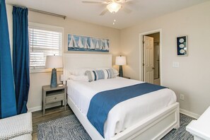A bedroom with a bed, two bedside tables, a ceiling fan, a window with blinds, and a painting on the wall.