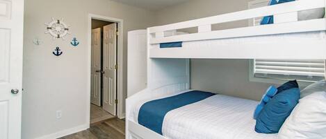 Ein Stockbett mit blauer Decke und Kissen, eine weiße Tür und ein Flur mit Holzboden.