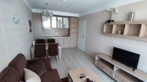 A modern living room with a brown sofa, a coffee table, and a flat-screen TV. There is a dining area with a table and chairs, and a kitchen with cabinets and a sink. - Luxuriöse Ferienwohnung im Harmony Suites Monte Carlo am Sonnenstrand (Sonnenstrand)