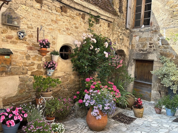 Un bâtiment en pierre au charme rustique, doté d’une petite fenêtre circulaire, d’une porte en bois et agrémenté de diverses plantes et fleurs en pot.