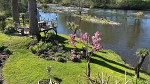 Udsigt over en flod med en terrasse og stole, omgivet af træer og grøn vegetation.