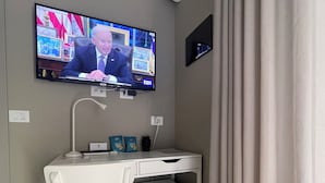 A hotel room with a white desk, a television mounted on the wall, a small refrigerator, and a patterned floor.