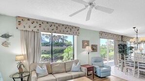 A spacious living room with a beige sofa, a blue armchair, a wooden coffee table, and a dining area with a white table and chairs.