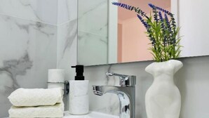 A bathroom with a marble wall, a white sink, a chrome faucet, a white vase with a plant, and a towel dispenser. - Corso Venezia - Suite (Venezia)