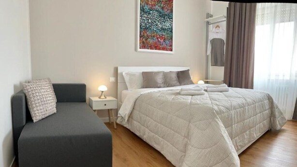 A modern hotel room with a bed, a sofa, a bedside table, and a painting on the wall. - Corso Venezia - Suite (Venezia)