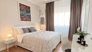 A modern bedroom with a large bed, bedside tables with lamps, a desk with a chair, and a large window with curtains. - Corso Venezia - Suite (Venezia)