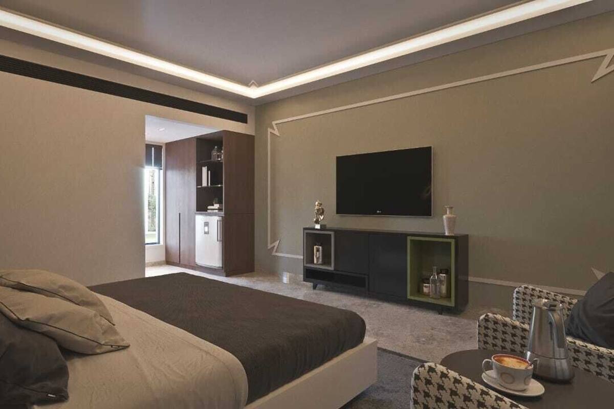 Una habitación de hotel moderna con una cama grande, un televisor de pantalla plana montado en la pared, un pequeño rincón de descanso con mesa y sillas, y una kitchenette al fondo.