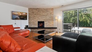 A modern living room with a stone fireplace, a large window with a view of greenery, and a red leather sofa.