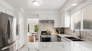 A modern kitchen with white cabinetry, a stainless steel refrigerator, a marble countertop, and a view of the outdoors through large windows.