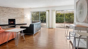 A spacious living room with a fireplace, a sofa, a dining area, and large windows offering a view of the outdoors.
