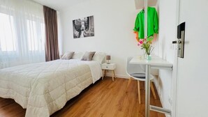 A bedroom with a bed, a nightstand, a desk, and a chair. There is a window with curtains, a wall-mounted TV, and a picture on the wall.