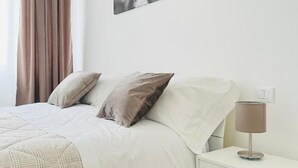 A neatly made bed with a white comforter and two pillows, a bedside table with a lamp, and a wall-mounted power outlet.