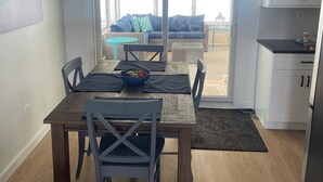 Zona pranzo con tavolo in legno, sedie blu e una ciotola di frutta. Sopra, una ventola a soffitto e una porta scorrevole in vetro che dà su un’area salotto esterna.
