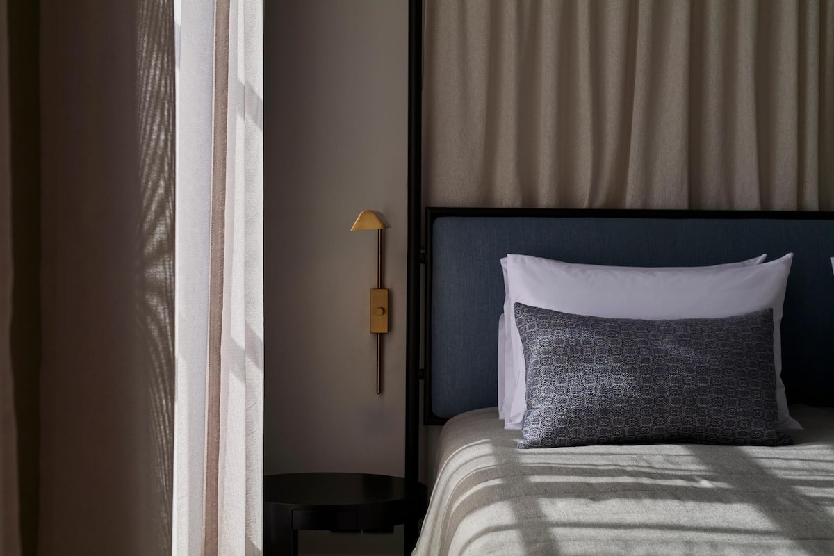 Una habitación de hotel con cama, mesita de noche, lámpara y ventana con cortinas.
