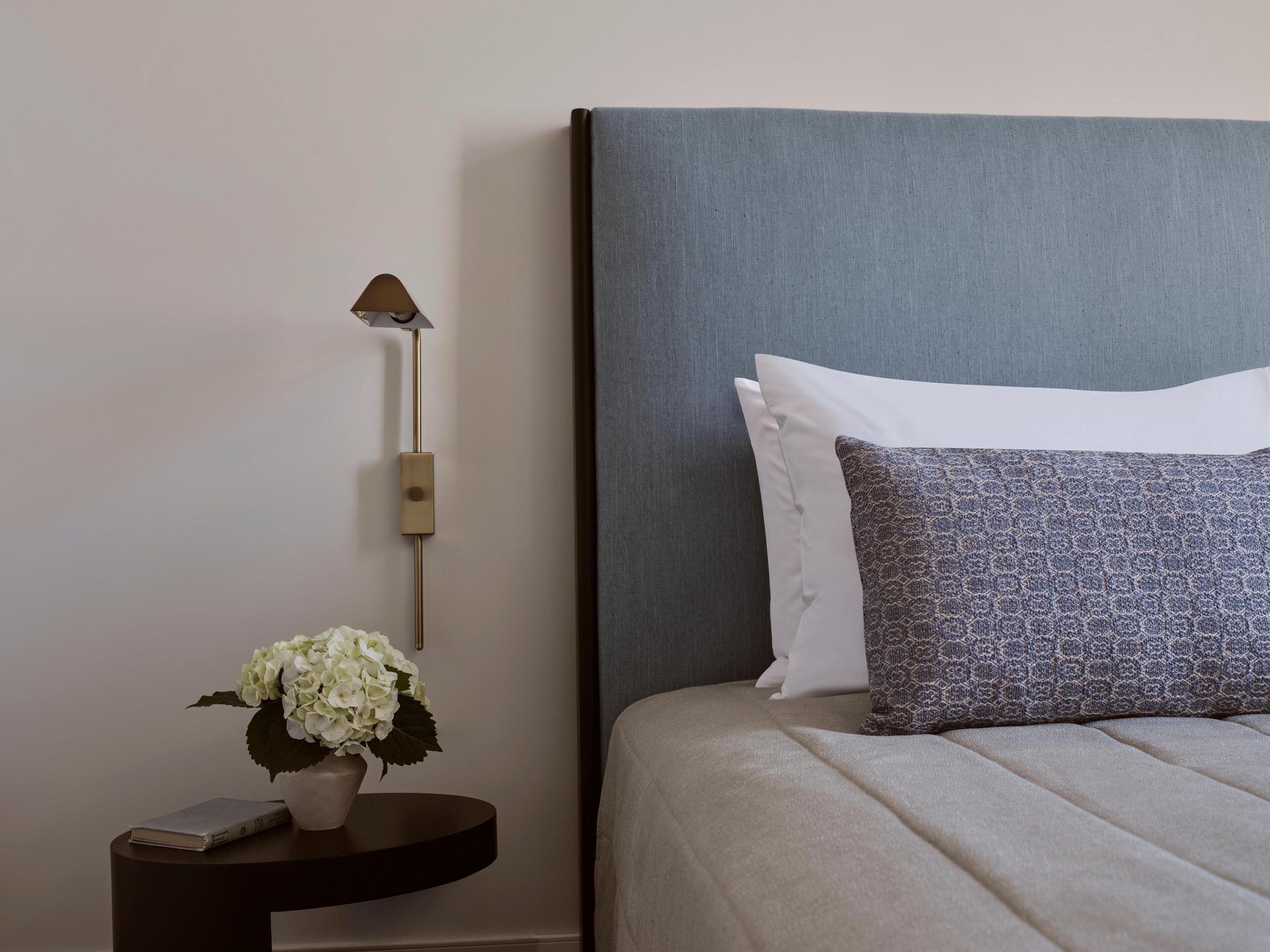 A neatly made bed with a grey headboard, white and patterned pillows, a small bedside table with a lamp, and a vase with white flowers.