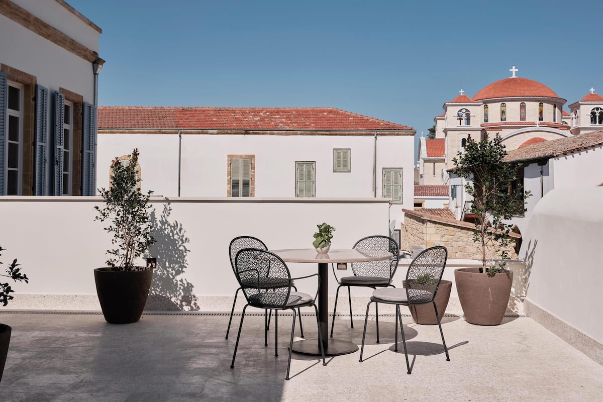 Una terraza en la azotea con una mesa y sillas, plantas en macetas y vistas a edificios tradicionales con tejados de tejas rojas.