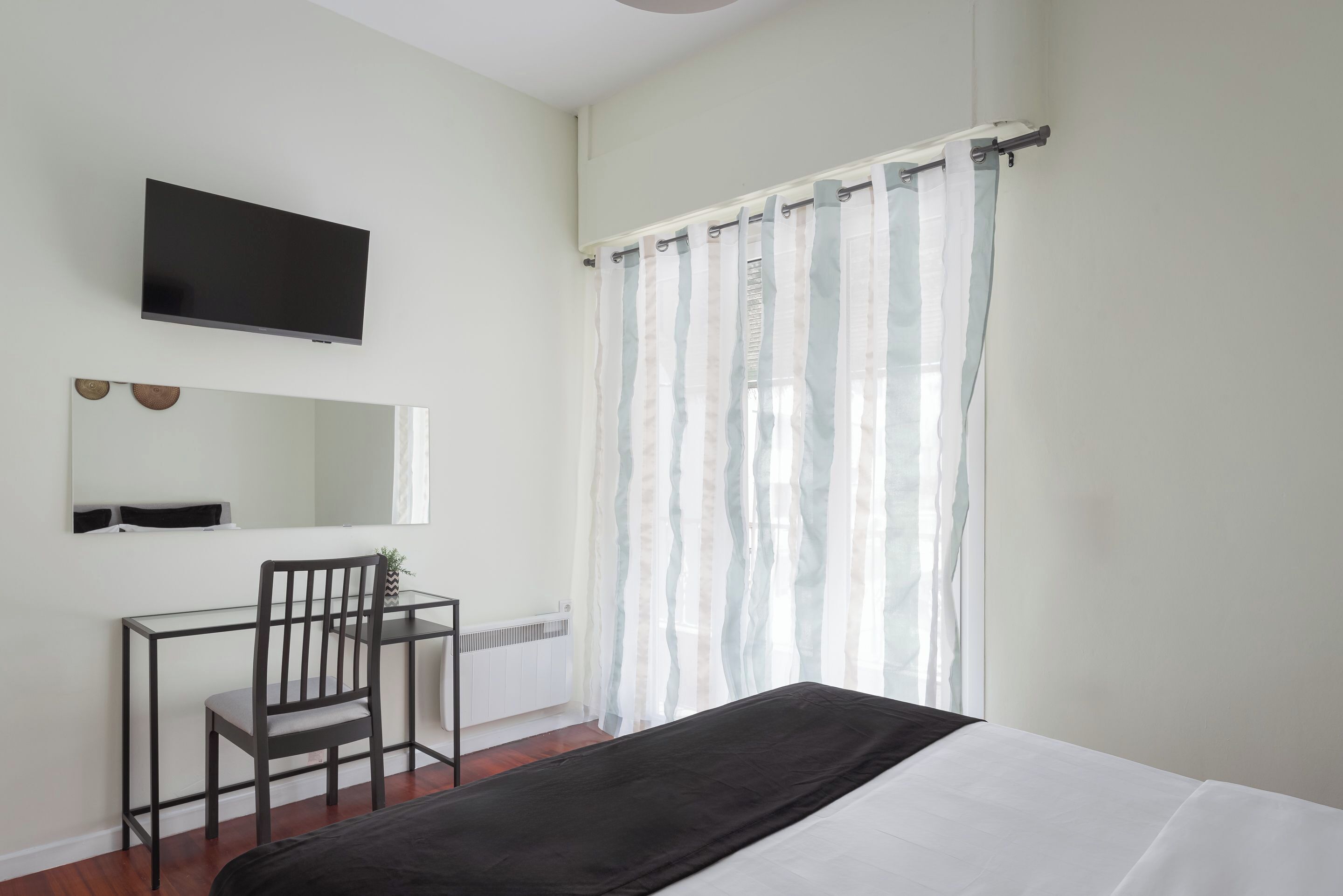 A bedroom with a bed, a desk with a chair, a TV mounted on the wall, and a window with sheer curtains.
