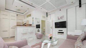 A modern living room with a glass coffee table, a sofa, a TV mounted on the wall, and a kitchen area in the background.