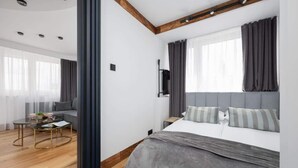 Un dormitorio moderno con una cama grande, televisor y sofá en una habitación luminosa con pisos de madera y ventanas amplias.