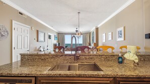 Una cocina con encimeras de granito, un lavamanos de acero inoxidable, sillas de madera y vistas al océano a través de la ventana.