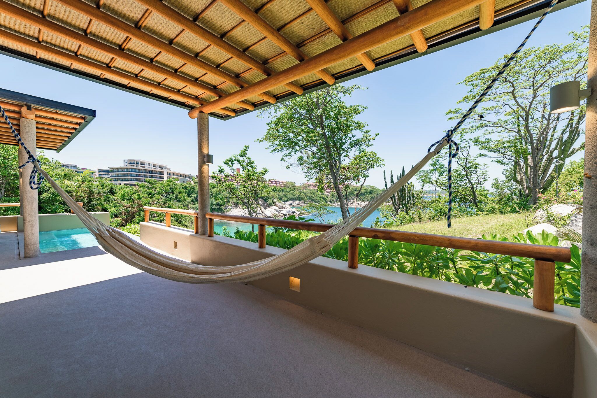 A hammock on a balcony overlooking a pool and lush greenery, with a view of distant buildings.
