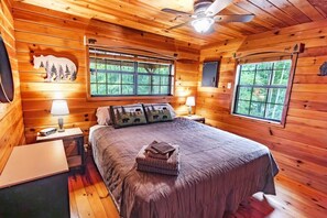 Une chambre à coucher dans un chalet en bois, avec un lit, deux lampes de chevet, un ventilateur de plafond et des fenêtres donnant sur la forêt.
