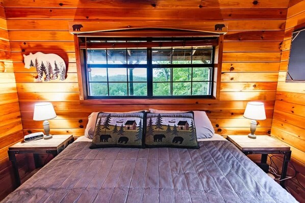Une chambre rustique en bois, avec un lit, deux tables de chevet, une fenêtre donnant sur la forêt et la silhouette d’un ours peinte sur le mur.