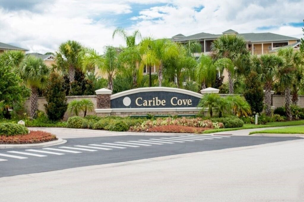 Schild für 'Caribe Cove' mit begrüntem Bereich davor, einschließlich Palmen und einer Straße mit Fußgängerüberwegen.