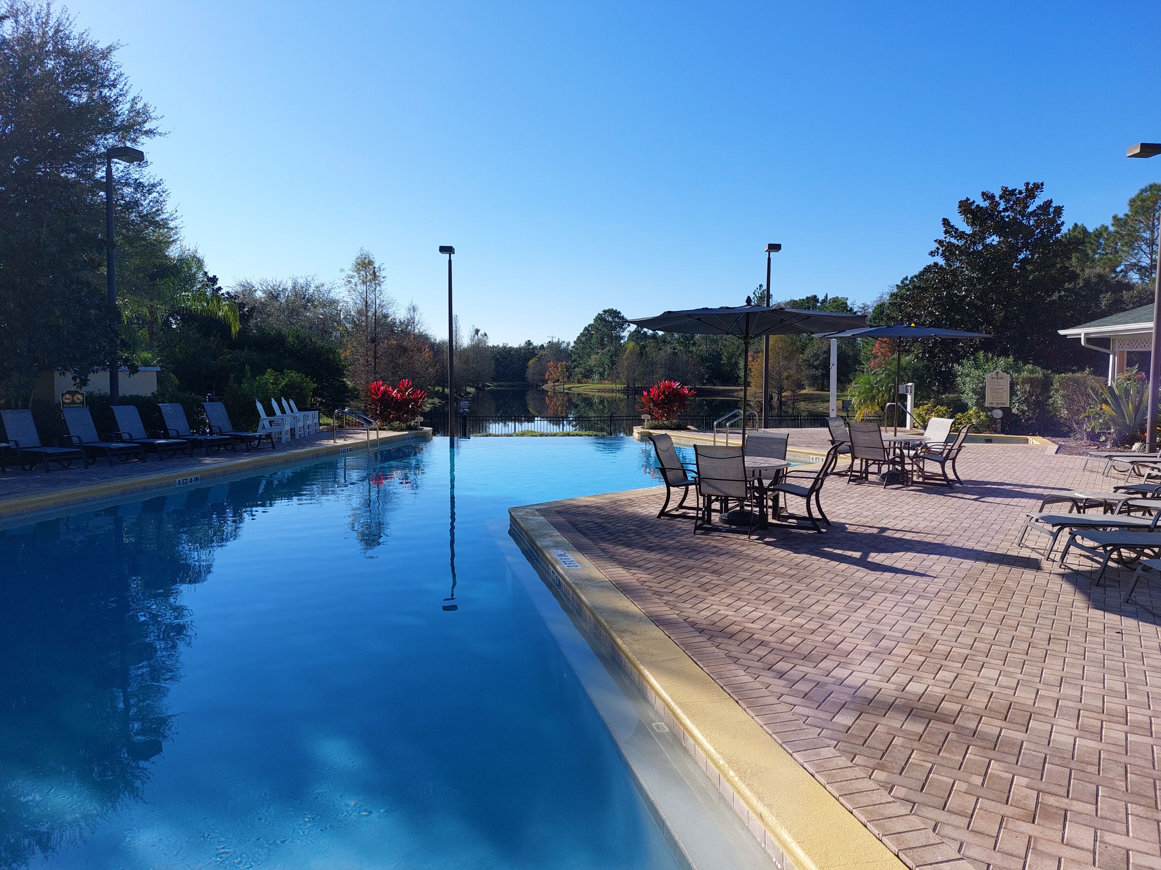 A swimming pool with lounge chairs and tables, surrounded by trees and a clear sky.